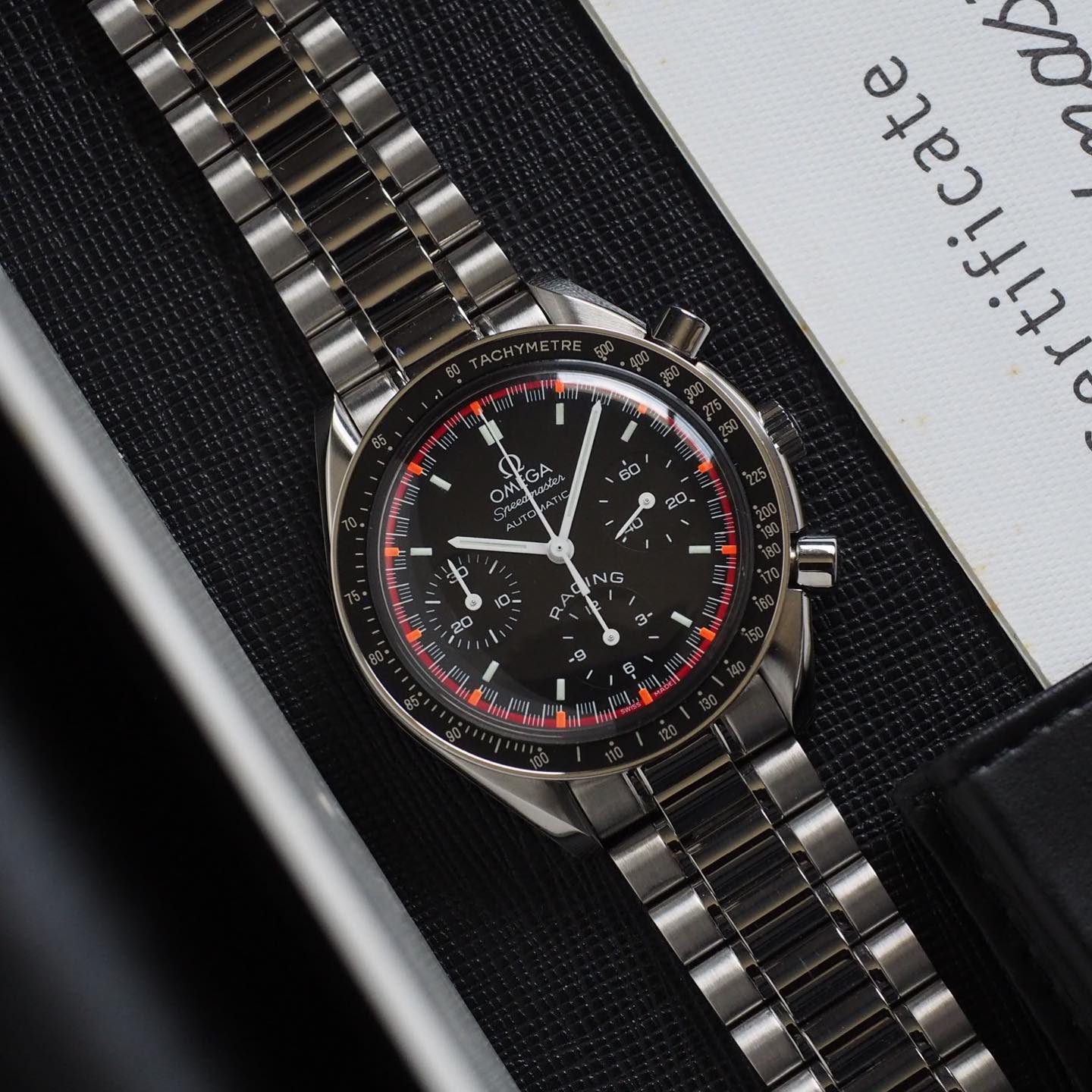 Schumacher dark dial with “Racing” print and checkered pattern. Orange and red outer ring along the seconds track. White hands. Painted indices with luminescent material. Omega Speedmaster Reduced 3518.50.00